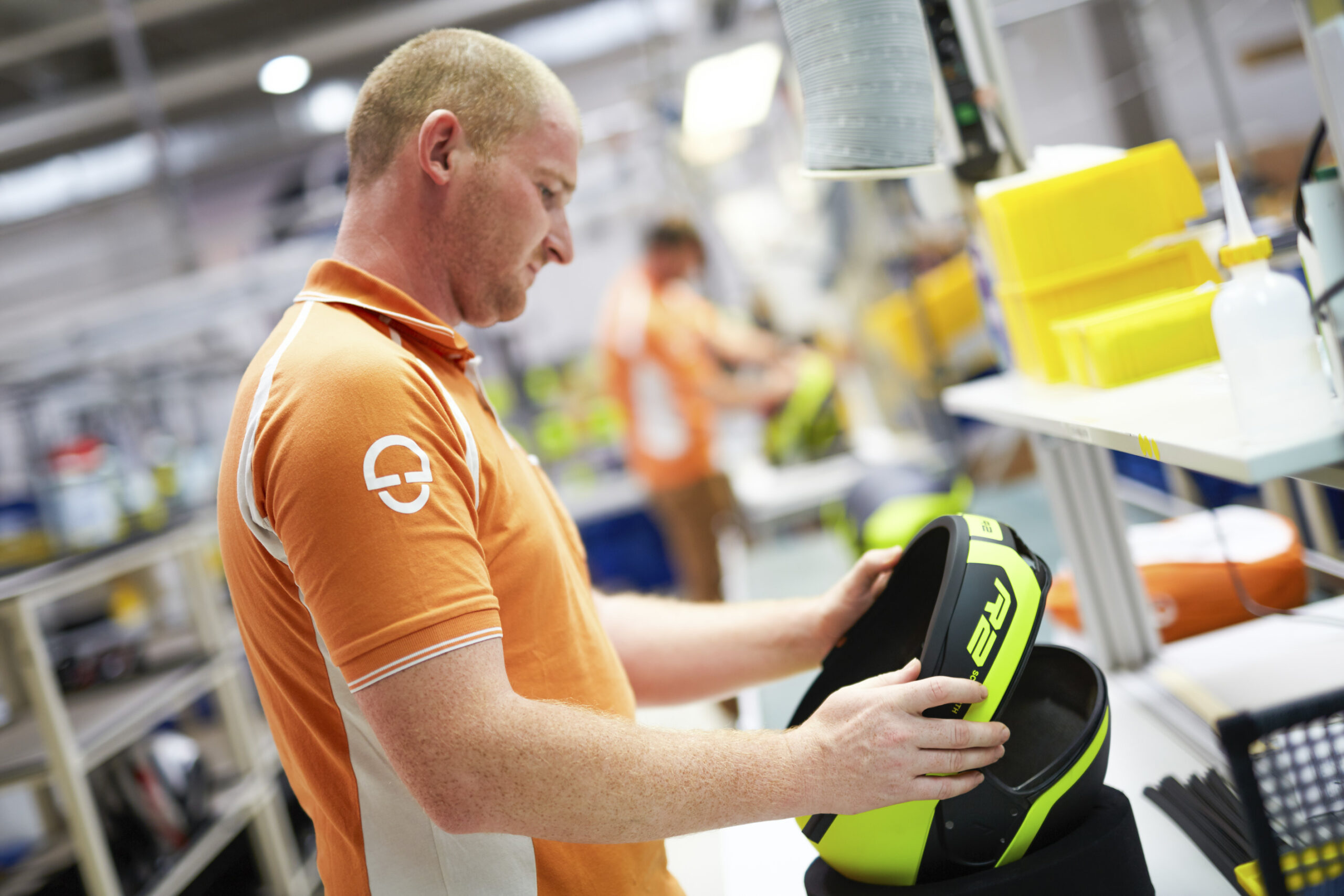 Mitarbeiter in orangefarbener Kleidung bearbeitet einen neongelben Schutzhelm in der Fertigung – Industriefotografie bei Schuberth