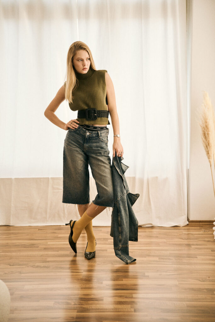 Startseite – Fotograf Magdeburg 18 Luisa kombiniert ein olivgrünes ärmelloses Stricktop mit weiter Denim-Bermuda, schwarzem Gürtel, gelben Socken und schwarzen Slingback-Pumps. Spiegel-, Türrahmen- und Ganzkörperaufnahmen vor hellen Vorhängen zeigen das Styling aus wechselnden Perspektiven im natürlichen Fensterlicht.